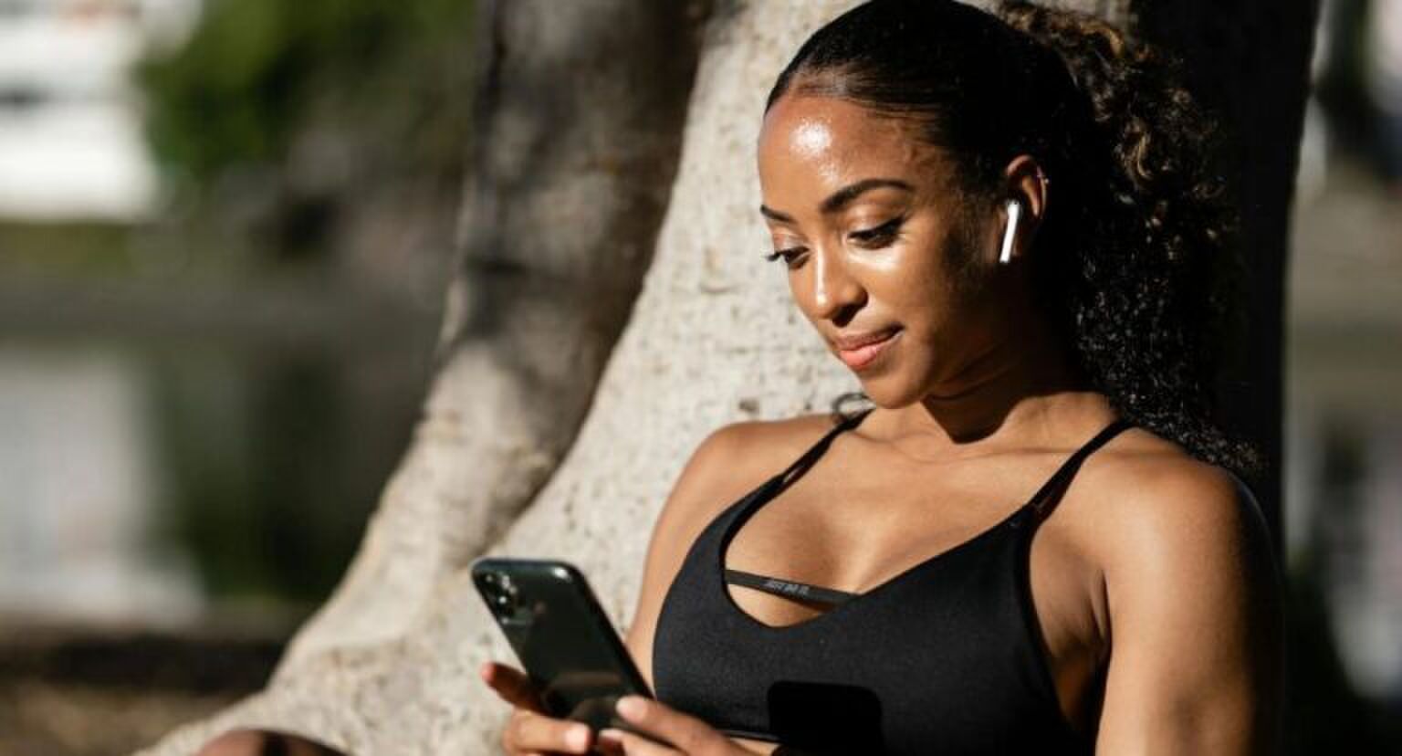 Vrouw met AirPods in die op haar telefoon zit terwijl ze tegen een boom aanleunt.