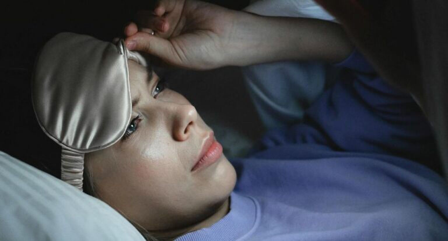 Een vrouw in bed die op haar telefoon kijkt terwijl ze haar slaapmasker omhoog tilt.
