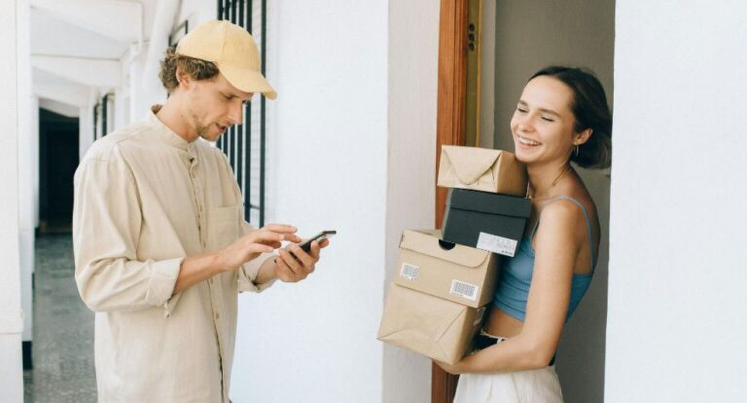 Een vrouw die veel pakketjes heeft ontvangen, en ze lachend vasthoudt. De postbode staat tegenover haar kijkend naar zijn telefoon.