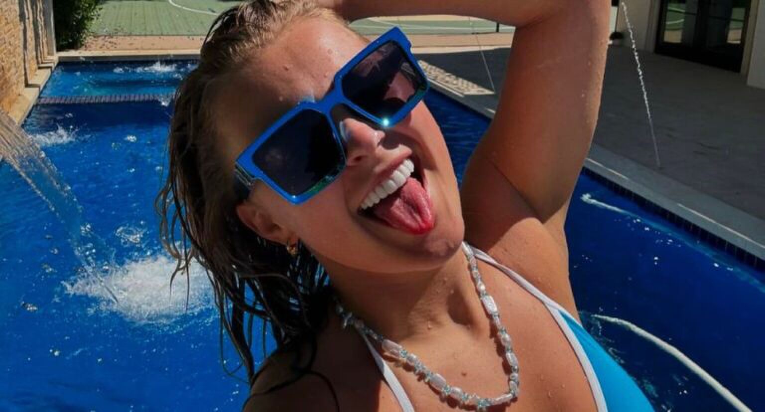 Selfie van JoJo Siwa waarin ze haar tong uitsteekt, een grote blauwe zonnebril op heeft en voor een zwembad staat.