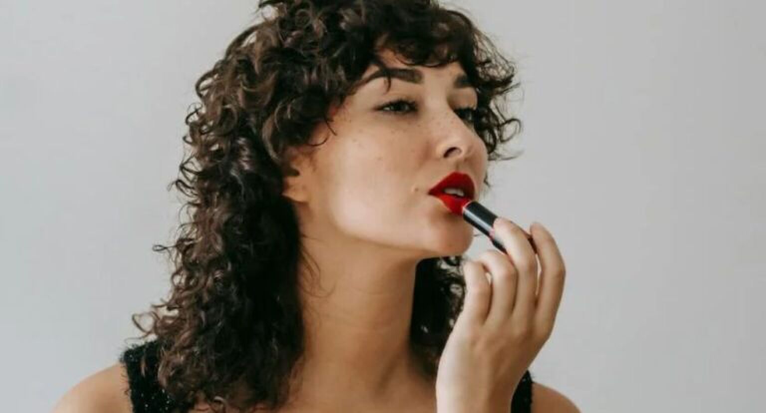 Een vrouw met krullen die rode lippenstift aanbrengt.