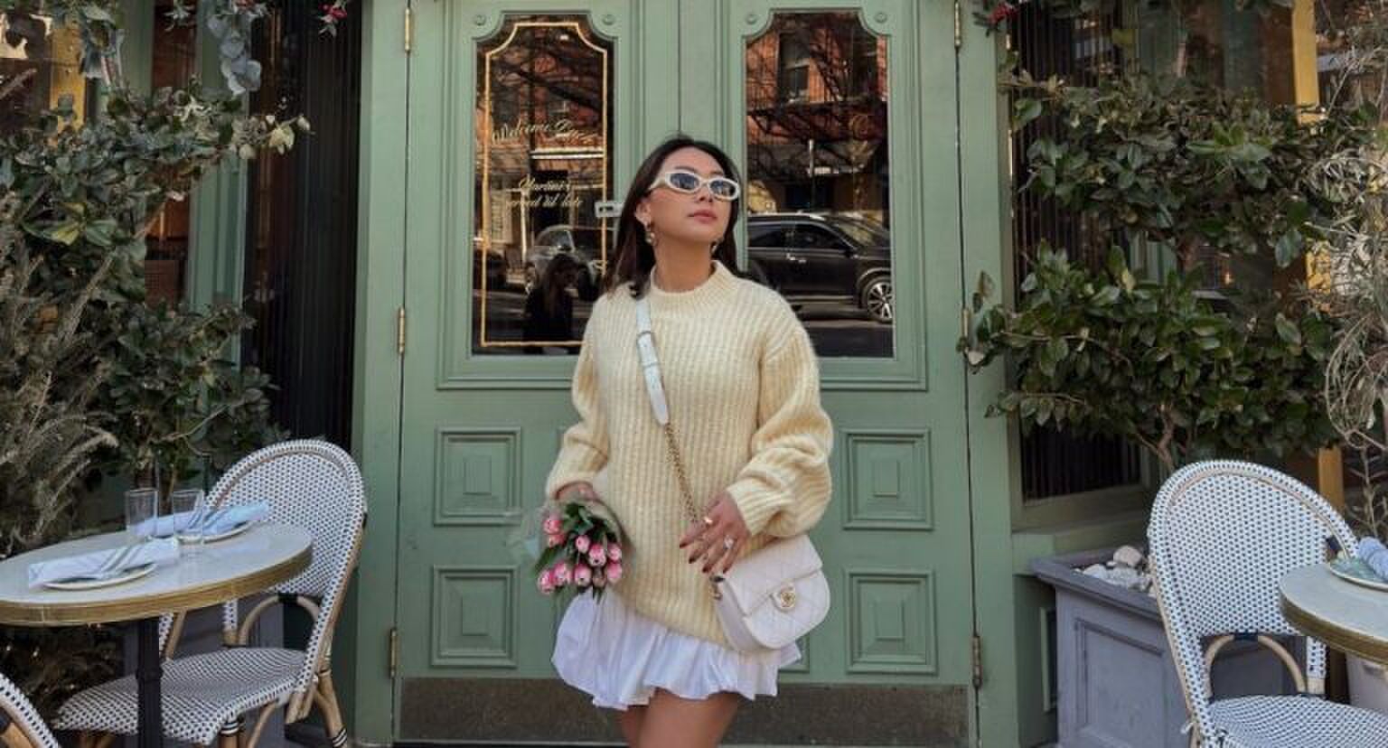 Een vrouw die voor een mooie groene deur staat, ze draagt een gele trui en een witte bubble skirt. In haar handen heeft ze een bosje tulpen.