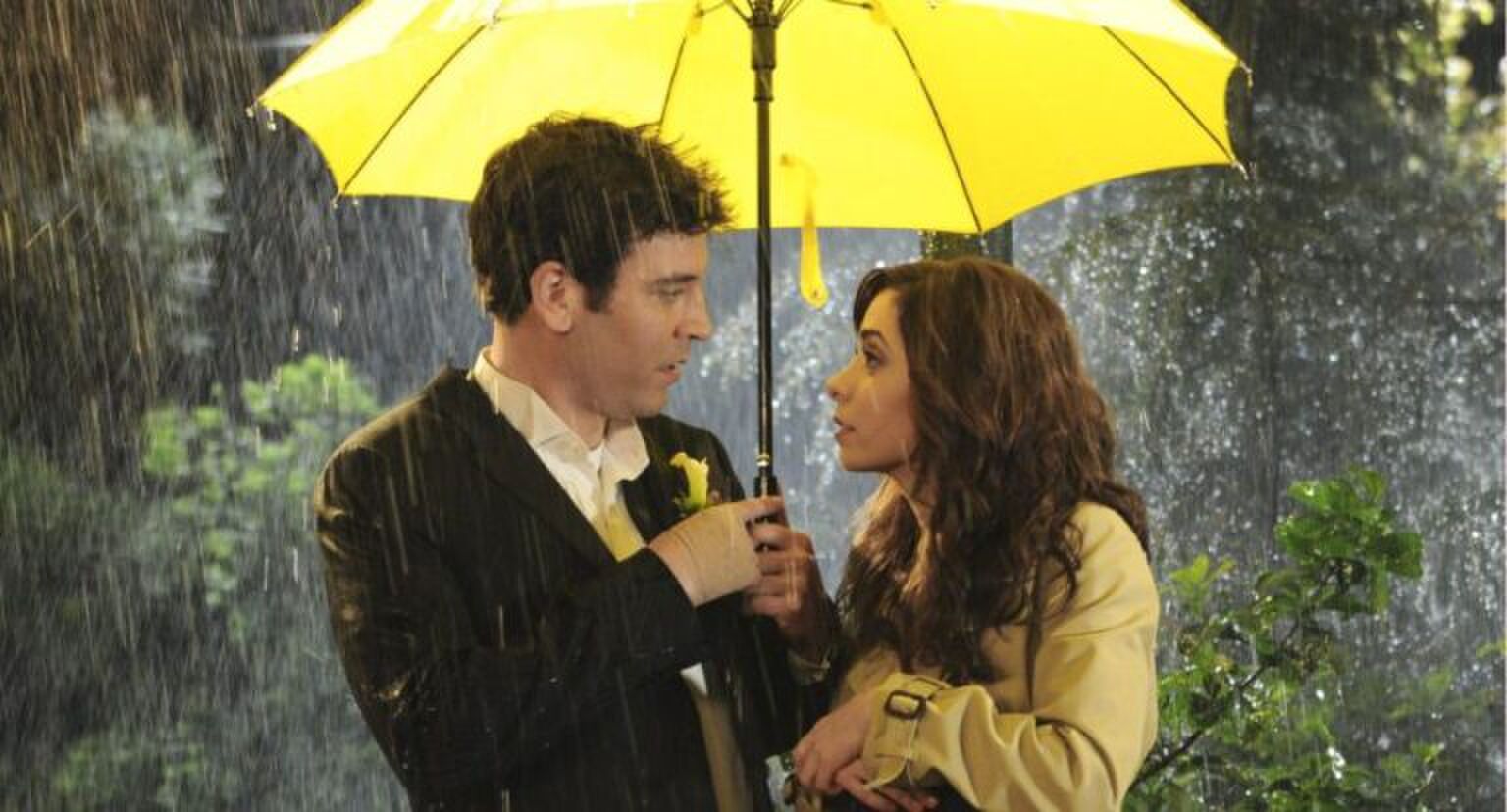 Man en vrouw kijken elkaar aan onder een gele paraplu in de stromende regen.