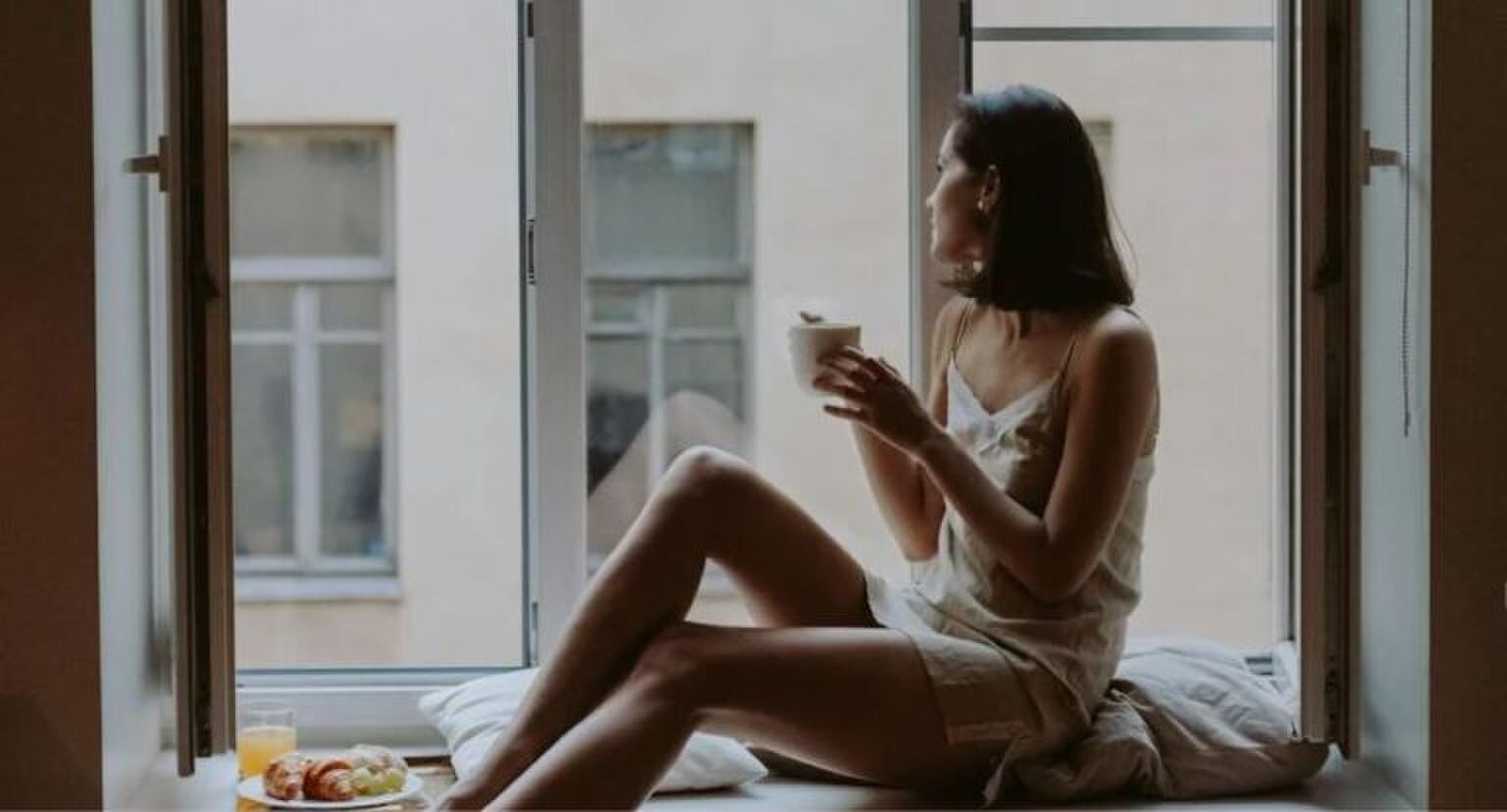 Vrouw kijkt uit een open raam met een kopje koffie in haar handen.