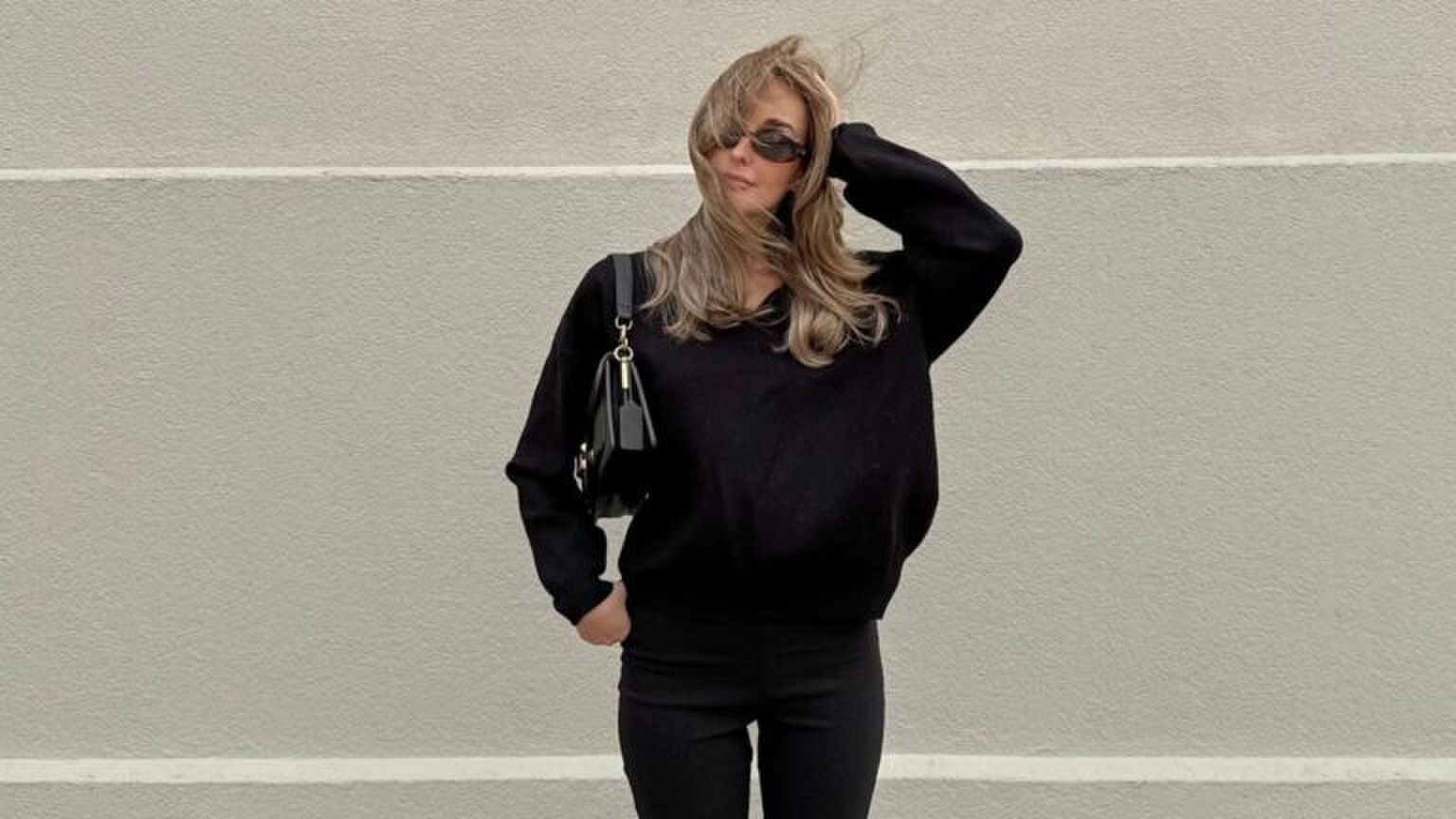 Een vrouw die poseert voor een witte muur in een compleet zwarte outfit.