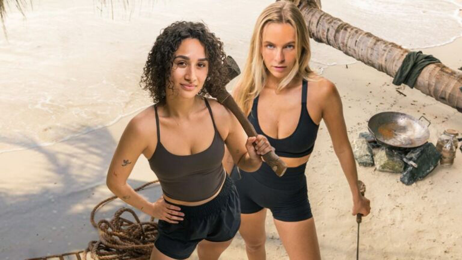 Twee vrouwen staan op een onbewoond eiland. De een heeft een bijl in haar hand, de ander een stok. Ze kijken stoer voor zich uit. Afbeelding uit Expeditie Robinson.