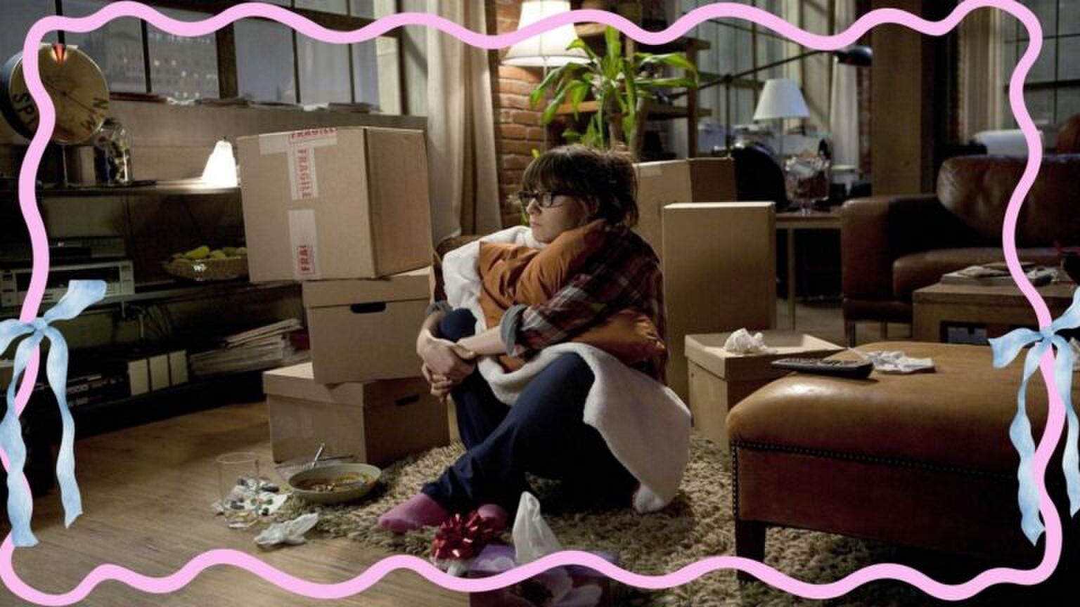 Vrouw kijkt sip. Ze is omring door verhuisdozen en heeft een bord met eten voor zich staan. Ze is net verhuisd. Sc&egrave;ne uit de serie: New Girl.