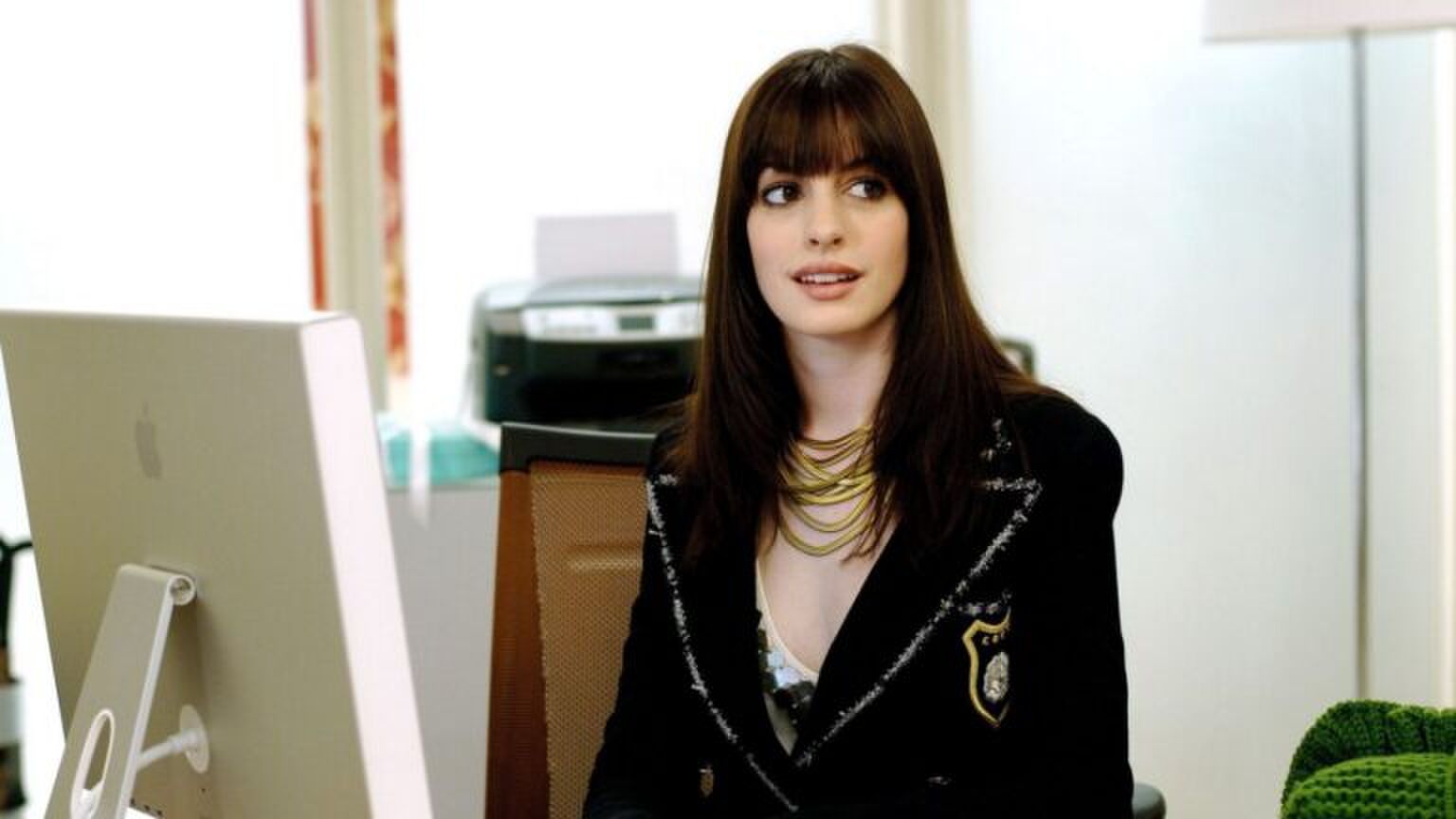 Emily van The Devil Wears Prada zit achter haar bureau.