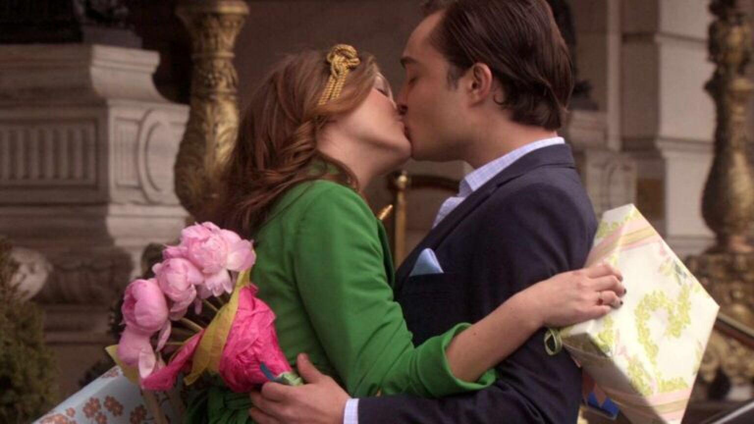 Leighton Meester en Ed Westwick als Blair en Chuck in de serie Gossip Girl. Ze geven elkaar een kus, terwijl ze een cadeau en een bos bloemen vasthouden.