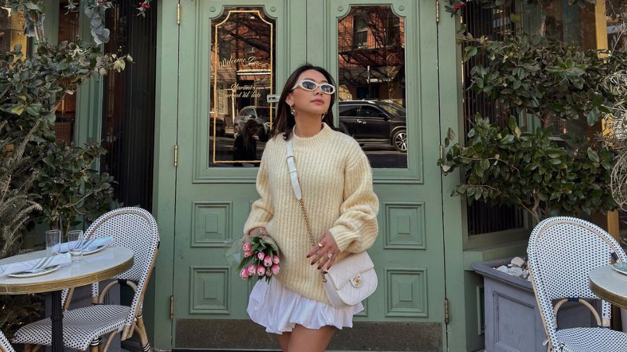 Een vrouw die voor een mooie groene deur staat, ze draagt een gele trui en een witte bubble skirt. In haar handen heeft ze een bosje tulpen.