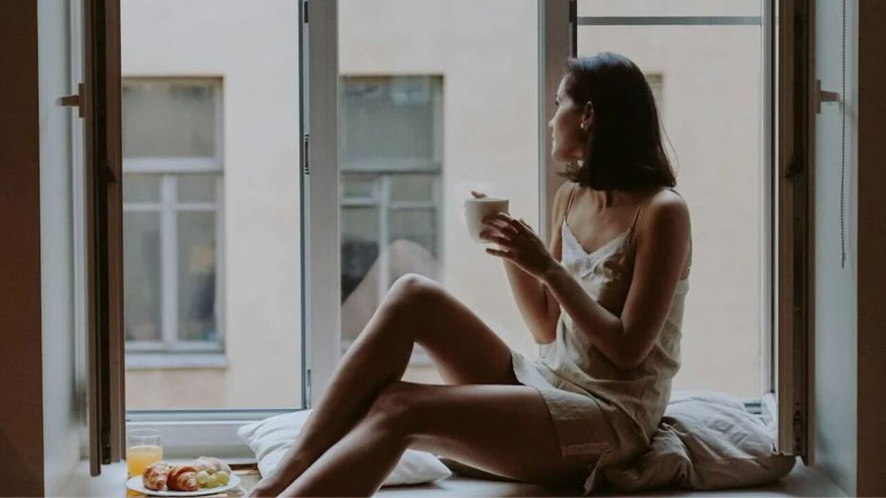 Vrouw kijkt uit een open raam met een kopje koffie in haar handen.