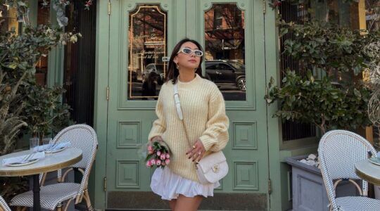 Een vrouw die voor een mooie groene deur staat, ze draagt een gele trui en een witte bubble skirt. In haar handen heeft ze een bosje tulpen.
