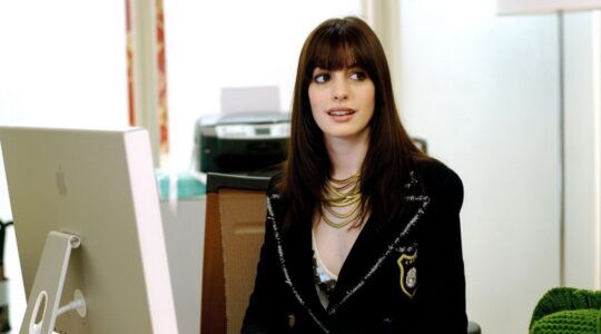 Emily van The Devil Wears Prada zit achter haar bureau.