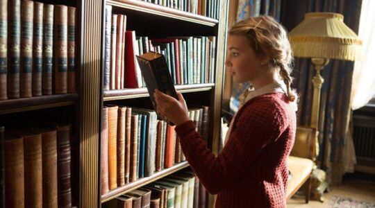 Een meisje staat in een bibliotheek. Ze pakt een boek uit een boekenkast. Ze kijkt geschokkeerd.