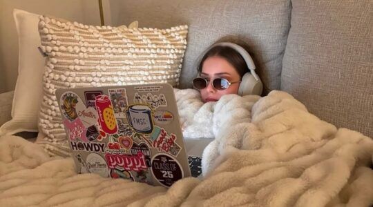 Vrouw in bed met haar laptop, bril en koptelefoon.