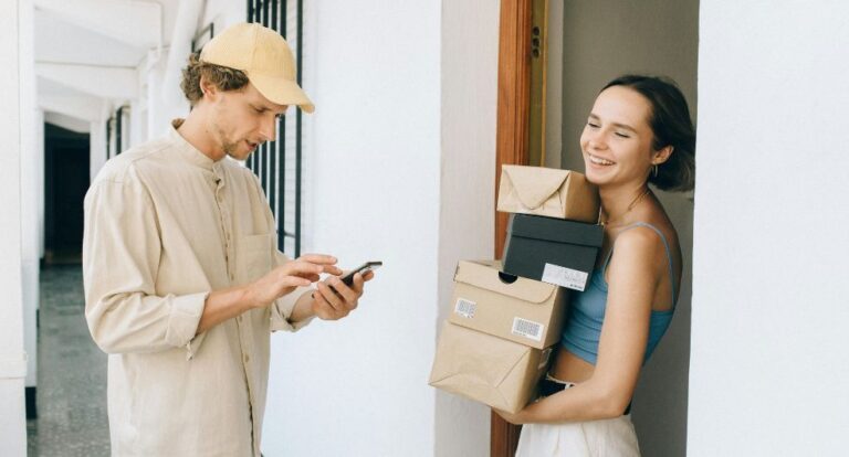 Een vrouw die veel pakketjes heeft ontvangen, en ze lachend vasthoudt. De postbode staat tegenover haar kijkend naar zijn telefoon.