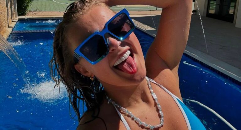 Selfie van JoJo Siwa waarin ze haar tong uitsteekt, een grote blauwe zonnebril op heeft en voor een zwembad staat.