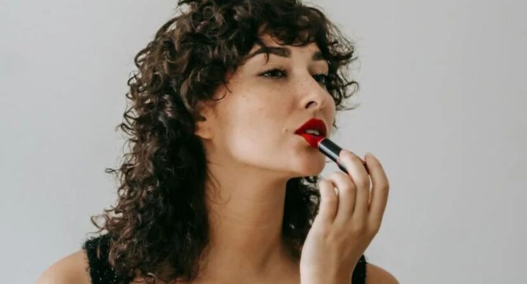Een vrouw met krullen die rode lippenstift aanbrengt.