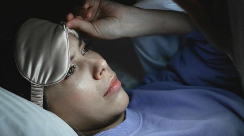 Een vrouw in bed die op haar telefoon kijkt terwijl ze haar slaapmasker omhoog tilt.