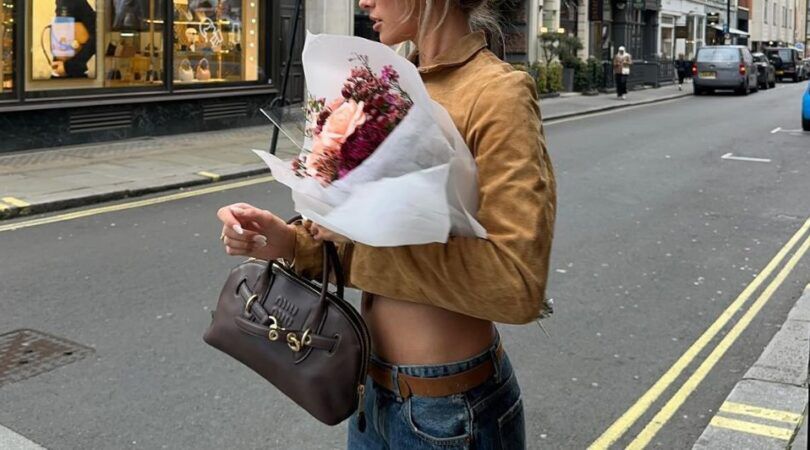 Meisje in jeans en okeren jas, met een bruine tas en een bos bloemen in haar armen.