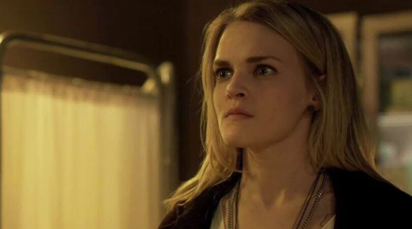 Een blonde vrouw kijkt met een serieuze blik langs de camera.