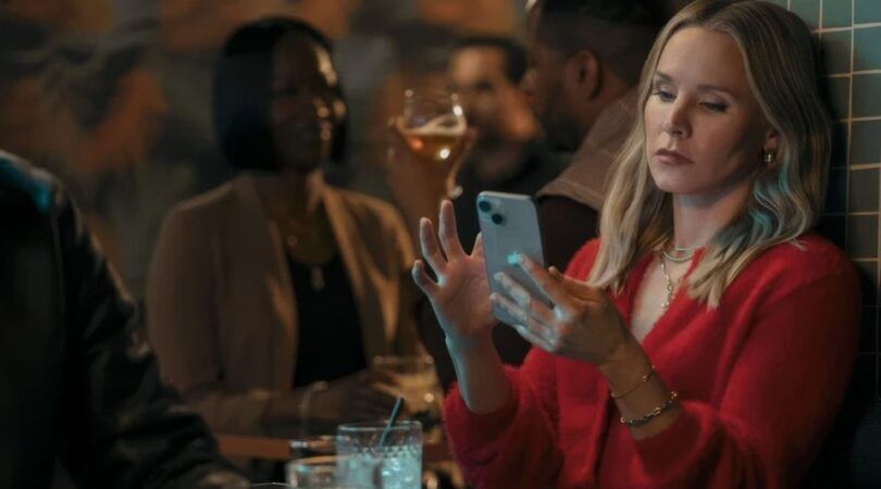 Kristen Bell kijkt vermoeid naar haar telefoon.