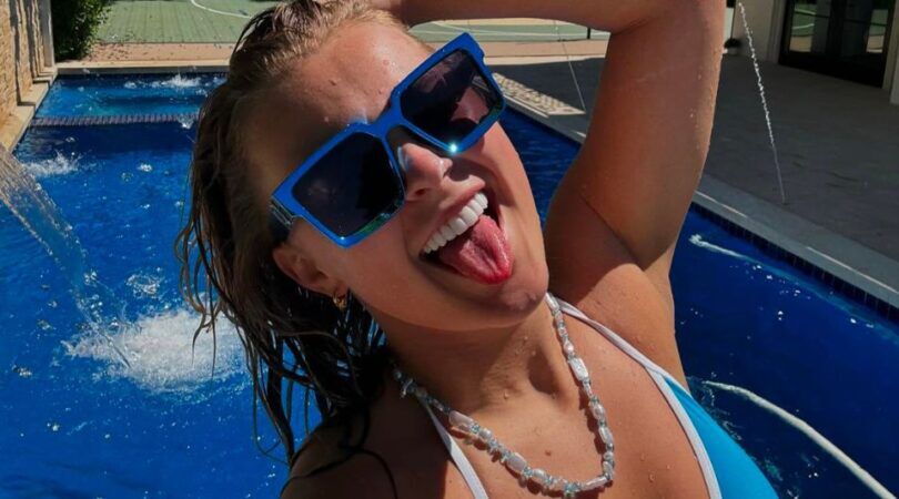 Selfie van JoJo Siwa waarin ze haar tong uitsteekt, een grote blauwe zonnebril op heeft en voor een zwembad staat.