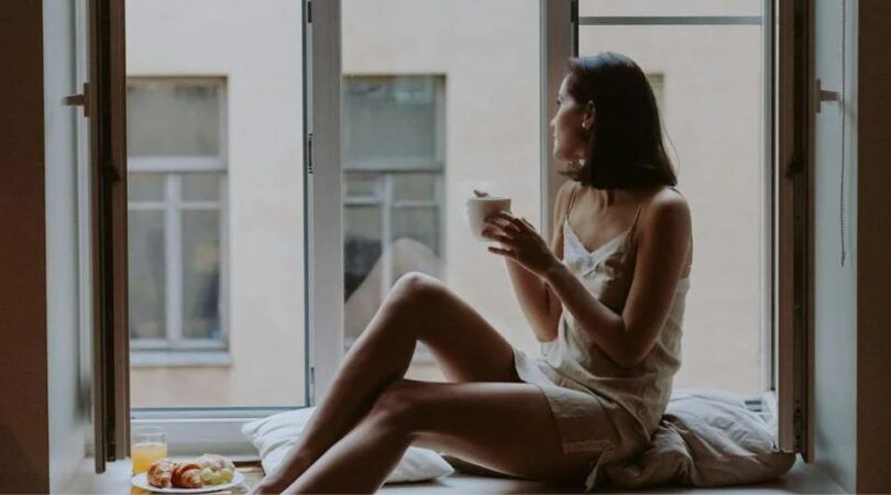 Vrouw kijkt uit een open raam met een kopje koffie in haar handen.