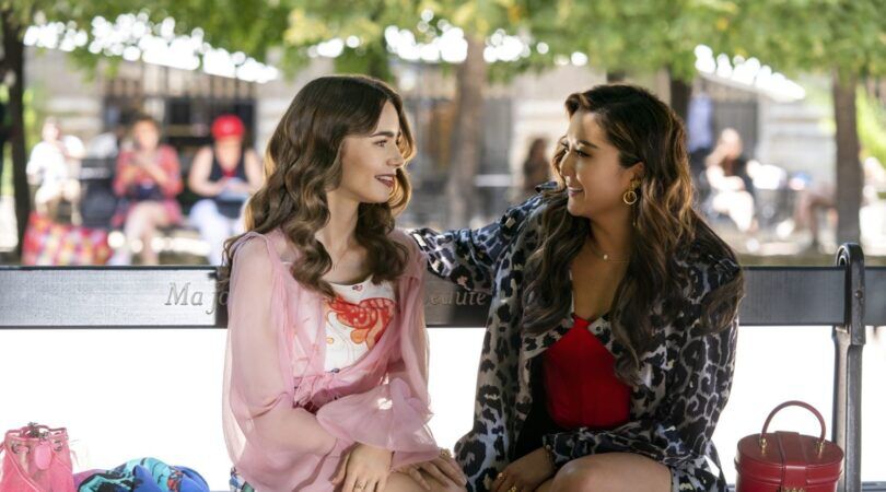 Emily en Mindy uit Emily in Paris zitten samen op een bankje.