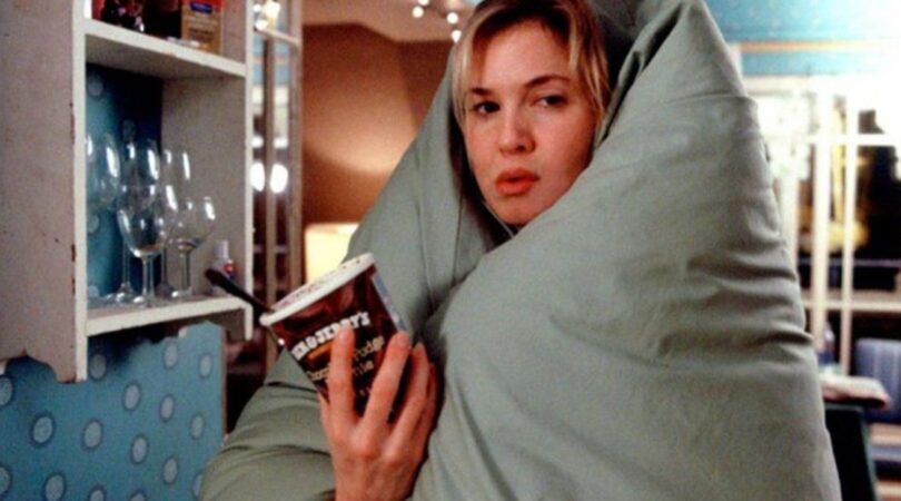 Luie vrouw, zit verwikkeld in een deken met een bak ijs in haar hand. Ze kijkt verveeld. Bridget Jones