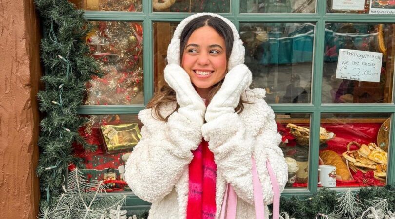 Vrouw kijkt lachend. Ze houdt haar handen in een V-vorm voor haar gezicht. Ze staat voor een kerstwinkel.