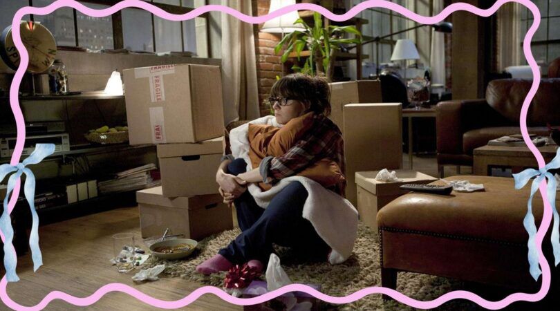 Vrouw kijkt sip. Ze is omring door verhuisdozen en heeft een bord met eten voor zich staan. Ze is net verhuisd. Sc&egrave;ne uit de serie: New Girl.