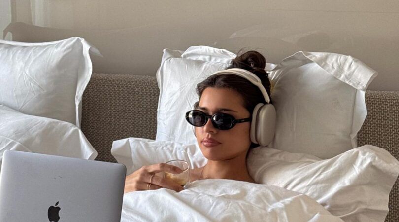 Meisje met koptelefoon en zonnebril op ligt in bed achter laptop.