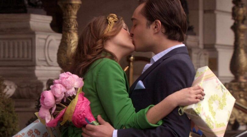 Leighton Meester en Ed Westwick als Blair en Chuck in de serie Gossip Girl. Ze geven elkaar een kus, terwijl ze een cadeau en een bos bloemen vasthouden.