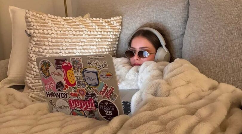 Vrouw in bed met haar laptop, bril en koptelefoon.